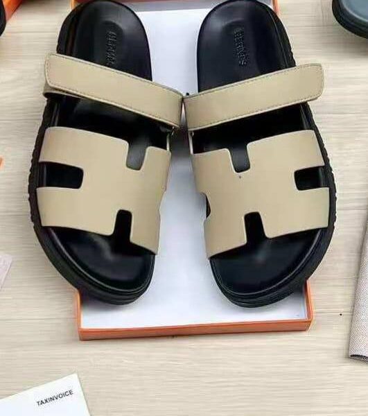 A pair of luxurious black and beige Hermes leather sandals, presented in an open orange box on a light wood surface.