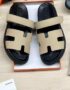 A pair of luxurious black and beige Hermes leather sandals, presented in an open orange box on a light wood surface.