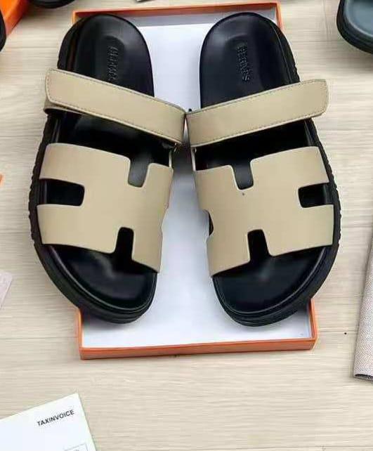 IMG-20260425-WA0045 A pair of luxurious black and beige Hermes leather sandals, presented in an open orange box on a light wood surface.