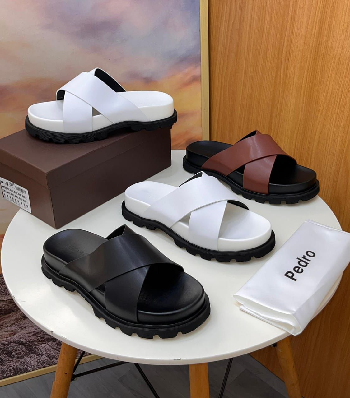 IMG-20260425-WA0048 A display of Pedro two-strap leather slide sandals in black, white, and brown on a stylish table setting, showcasing their sleek design and luxurious appeal.