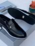 A pair of black leather loafers with white sole and silver logo on a shoe box with a plant in the background.