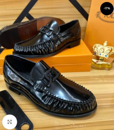 A pair of black leather tassel loafers with fringed details, displayed on a wooden surface with a shoe box and accessories in the background.