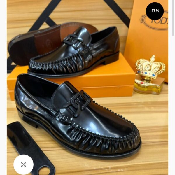 A pair of black leather tassel loafers with fringed details, displayed on a wooden surface with a shoe box and accessories in the background.