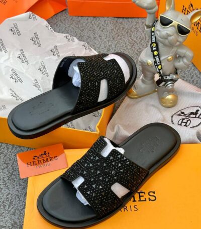 A pair of black studded Hermes Paris sandals on a yellow box with the brand's logo, accompanied by a small statue and packaging materials.