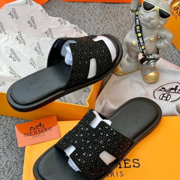 A pair of black studded Hermes Paris sandals on a yellow box with the brand's logo, accompanied by a small statue and packaging materials.