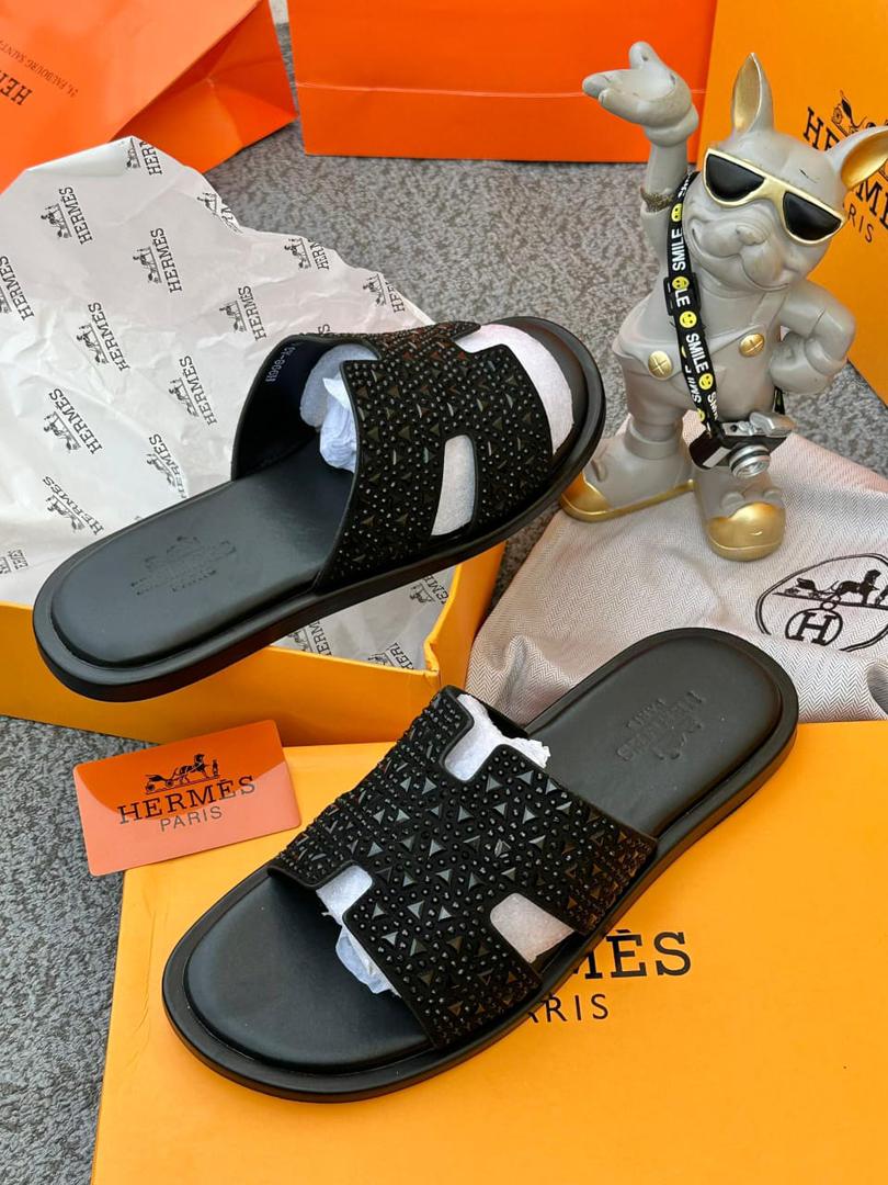 IMG-20260425-WA0052 A pair of black studded Hermes Paris sandals on a yellow box with the brand's logo, accompanied by a small statue and packaging materials.