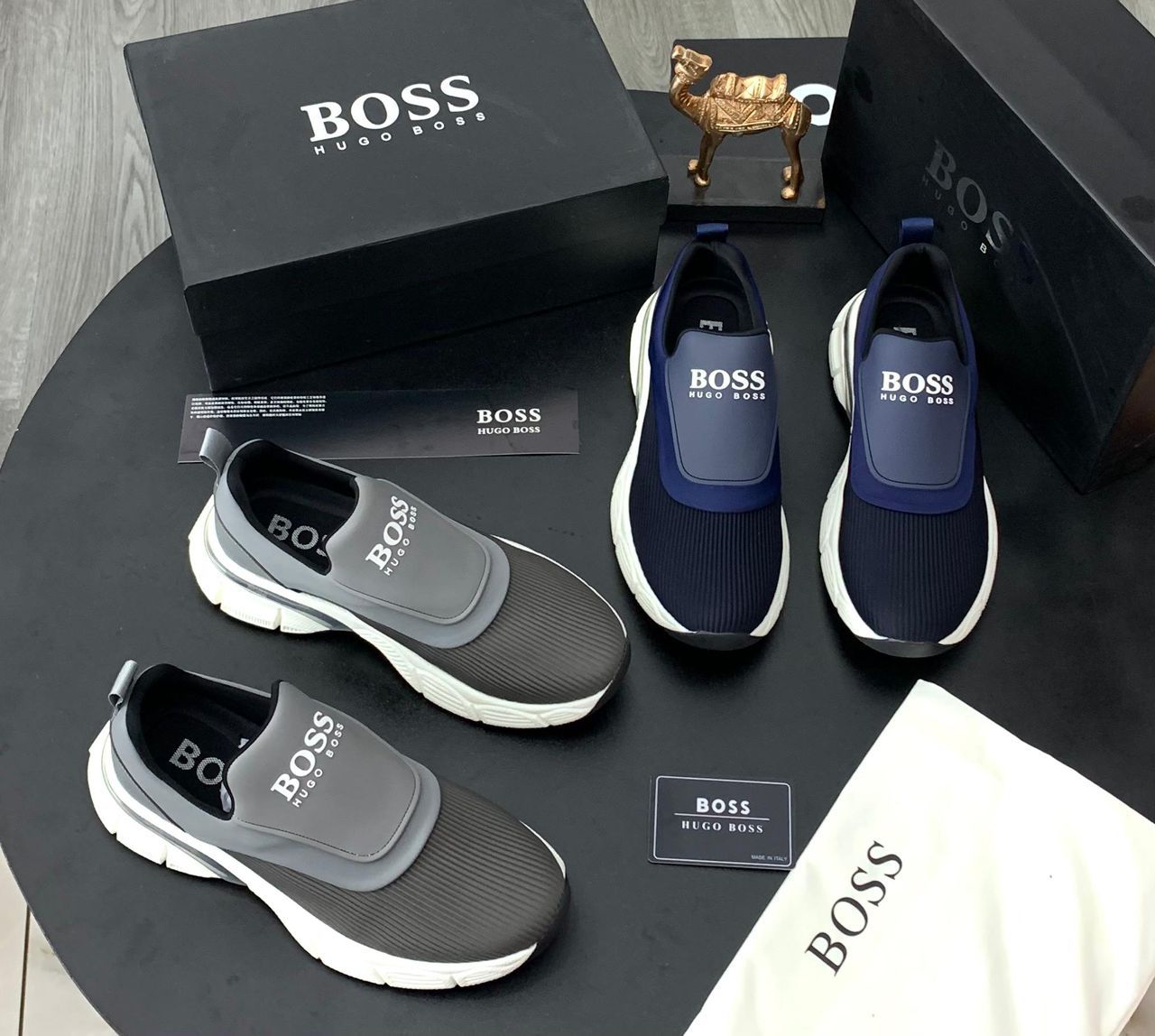 IMG-20260425-WA0053 A display of Hugo Boss men's slip-on sneakers in navy blue and grey, accompanied by their original boxes and packaging.