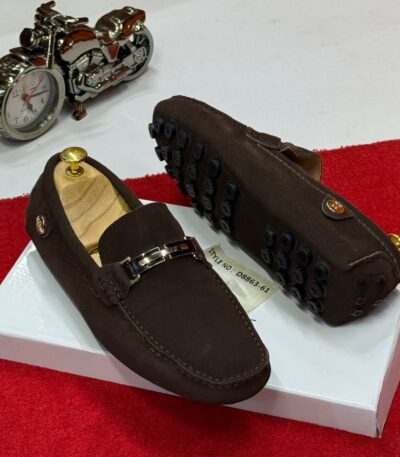 A pair of brown leather loafers with gold buckle and black studs on a white box, with a motorcycle clock in the background.
