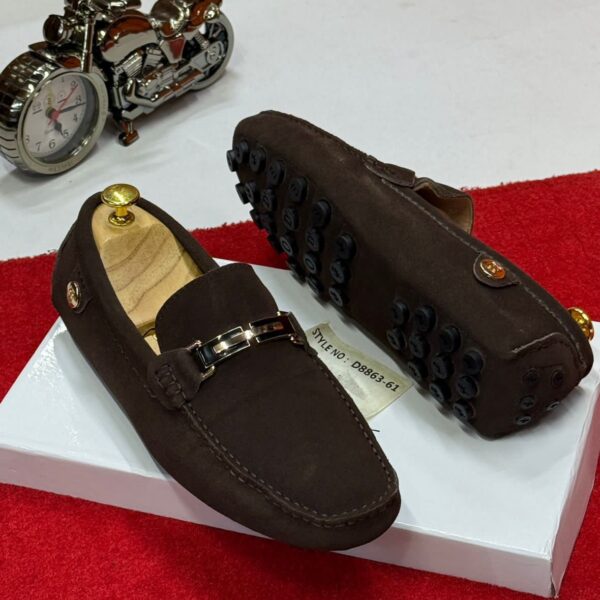 A pair of brown leather loafers with gold buckle and black studs on a white box, with a motorcycle clock in the background.