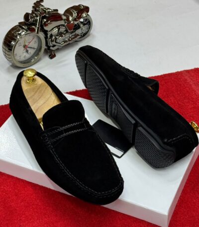 A pair of black suede loafers on a white box with a red carpet background, showcasing the shoes' sleek design and luxurious suede material.
