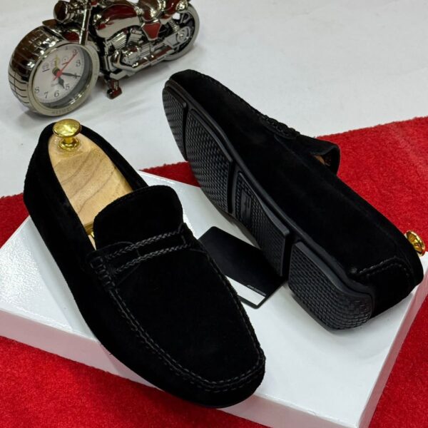 A pair of black suede loafers on a white box with a red carpet background, showcasing the shoes' sleek design and luxurious suede material.