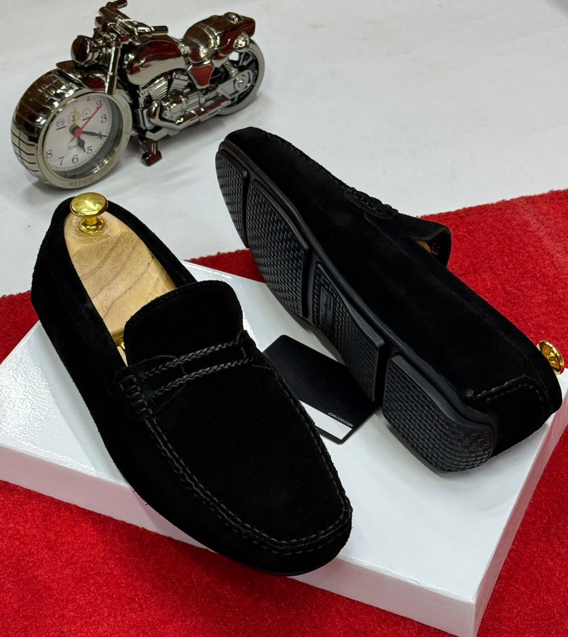 IMG-20260425-WA0055 A pair of black suede loafers on a white box with a red carpet background, showcasing the shoes' sleek design and luxurious suede material.