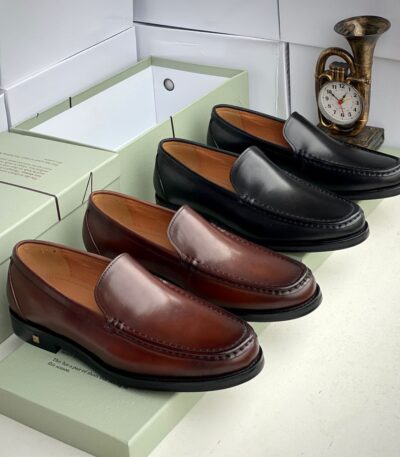 A pair of rich brown and sleek black leather loafers displayed on green shoe boxes, with a vintage brass French horn clock in the background, showcasing luxury and sophistication.