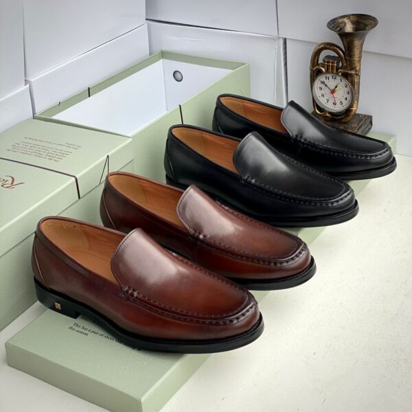 A pair of rich brown and sleek black leather loafers displayed on green shoe boxes, with a vintage brass French horn clock in the background, showcasing luxury and sophistication.
