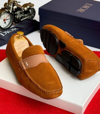 A pair of tan suede loafers with a signature buckle detail, displayed on a white box with a Dior box and accessories in the background.