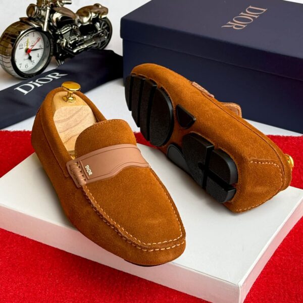 A pair of tan suede loafers with a signature buckle detail, displayed on a white box with a Dior box and accessories in the background.