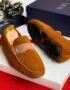 A pair of tan suede loafers with a signature buckle detail, displayed on a white box with a Dior box and accessories in the background.