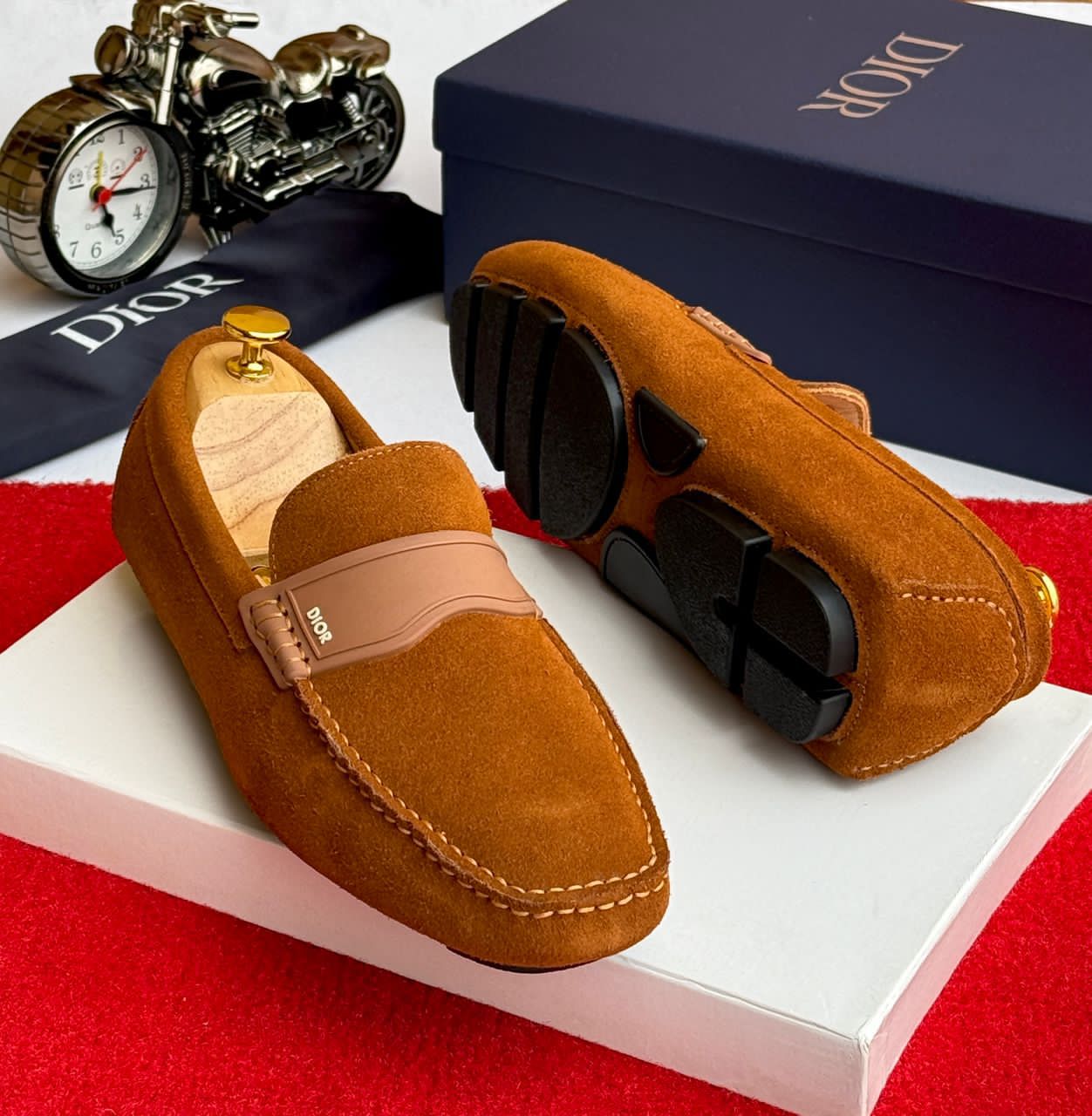 IMG-20260425-WA0059 A pair of tan suede loafers with a signature buckle detail, displayed on a white box with a Dior box and accessories in the background.