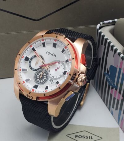 A rose gold Fossil watch with a black strap and white dial, displayed on a white surface with the Fossil logo and packaging in the background.