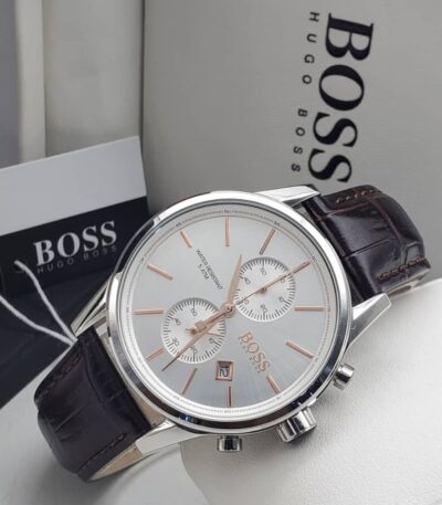 A close-up of a Hugo Boss men's watch with a silver-toned case, white dial, and brown leather strap, displayed on a white surface with the brand's packaging in the background.