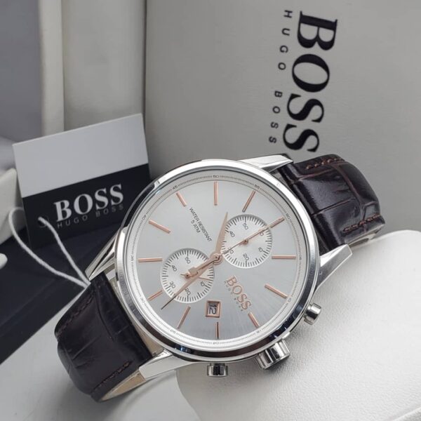 A close-up of a Hugo Boss men's watch with a silver-toned case, white dial, and brown leather strap, displayed on a white surface with the brand's packaging in the background.