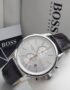 A close-up of a Hugo Boss men's watch with a silver-toned case, white dial, and brown leather strap, displayed on a white surface with the brand's packaging in the background.