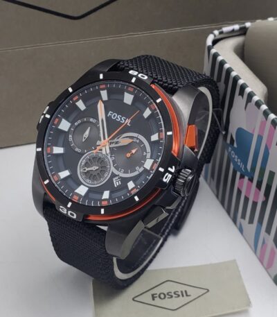 A black and orange Fossil chronograph watch with a black strap, displayed on a white surface with a colorful box and tag in the background.