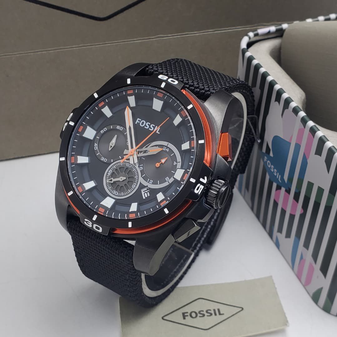 IMG-20260612-WA0009 A black and orange Fossil chronograph watch with a black strap, displayed on a white surface with a colorful box and tag in the background.