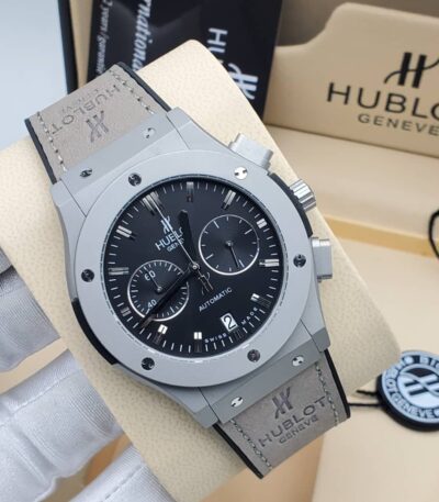 A close-up view of the Hublot Big Bang Automatic watch with a grey case and black dial, displayed on a beige stand with the Hublot logo in the background.