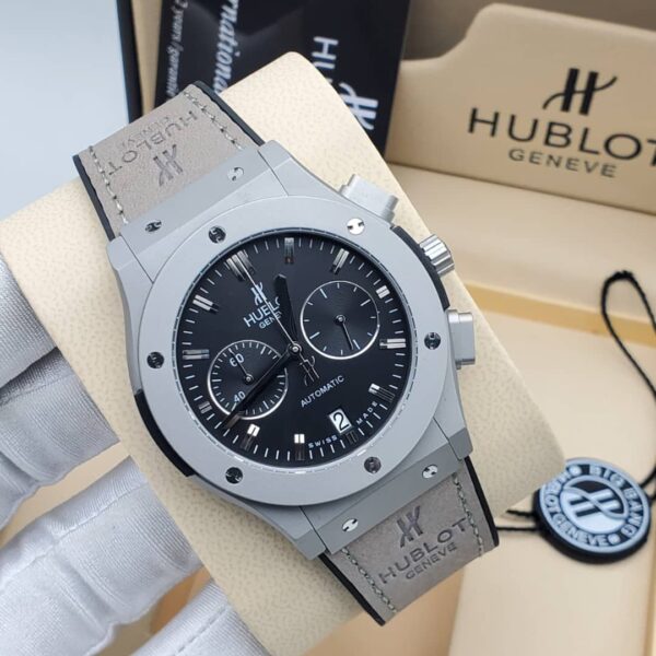 A close-up view of the Hublot Big Bang Automatic watch with a grey case and black dial, displayed on a beige stand with the Hublot logo in the background.