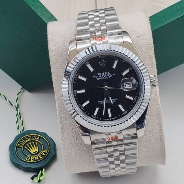 A luxurious Rolex Oyster Perpetual Datejust men's watch with a black dial and silver band, displayed on a white cushion with a green box and Rolex logo in the background.