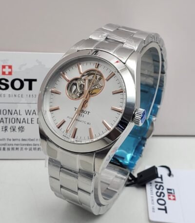 A sleek Tissot watch with a silver band and white face, featuring a visible mechanical movement, displayed on a white background with its box and tag.
