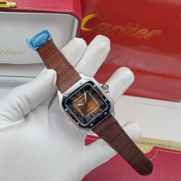 A luxurious Cartier watch with a brown leather strap and silver case, featuring a brown dial with Roman numerals, held by a gloved hand with the Cartier box in the background.