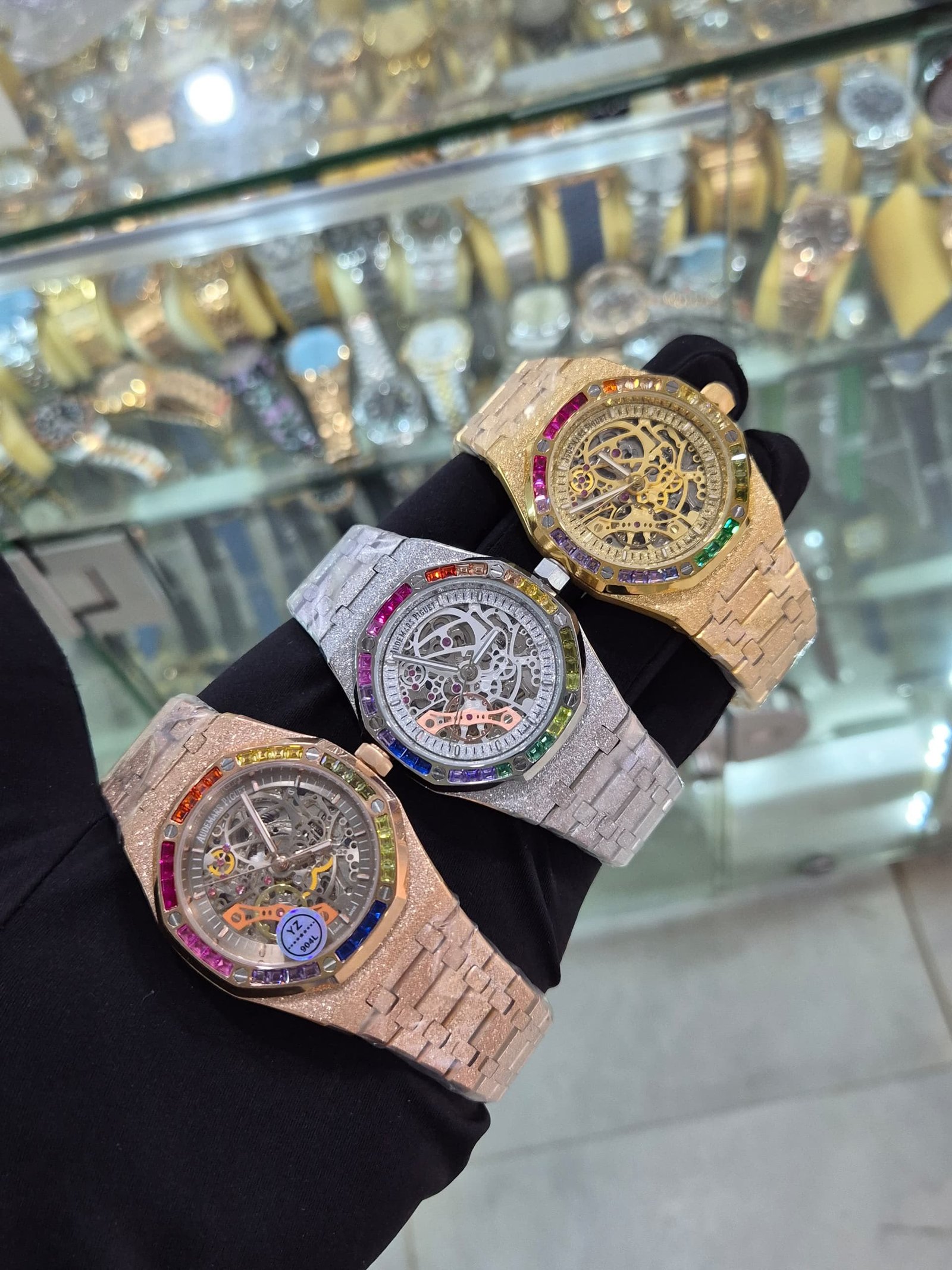 root A hand showcasing three luxurious watches with rainbow gemstone bezels and intricate mechanical details.