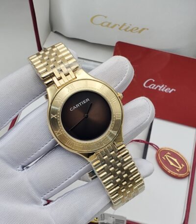 A gold Cartier watch with a black dial and Roman numerals, displayed on a gloved hand with the brand's packaging in the background.