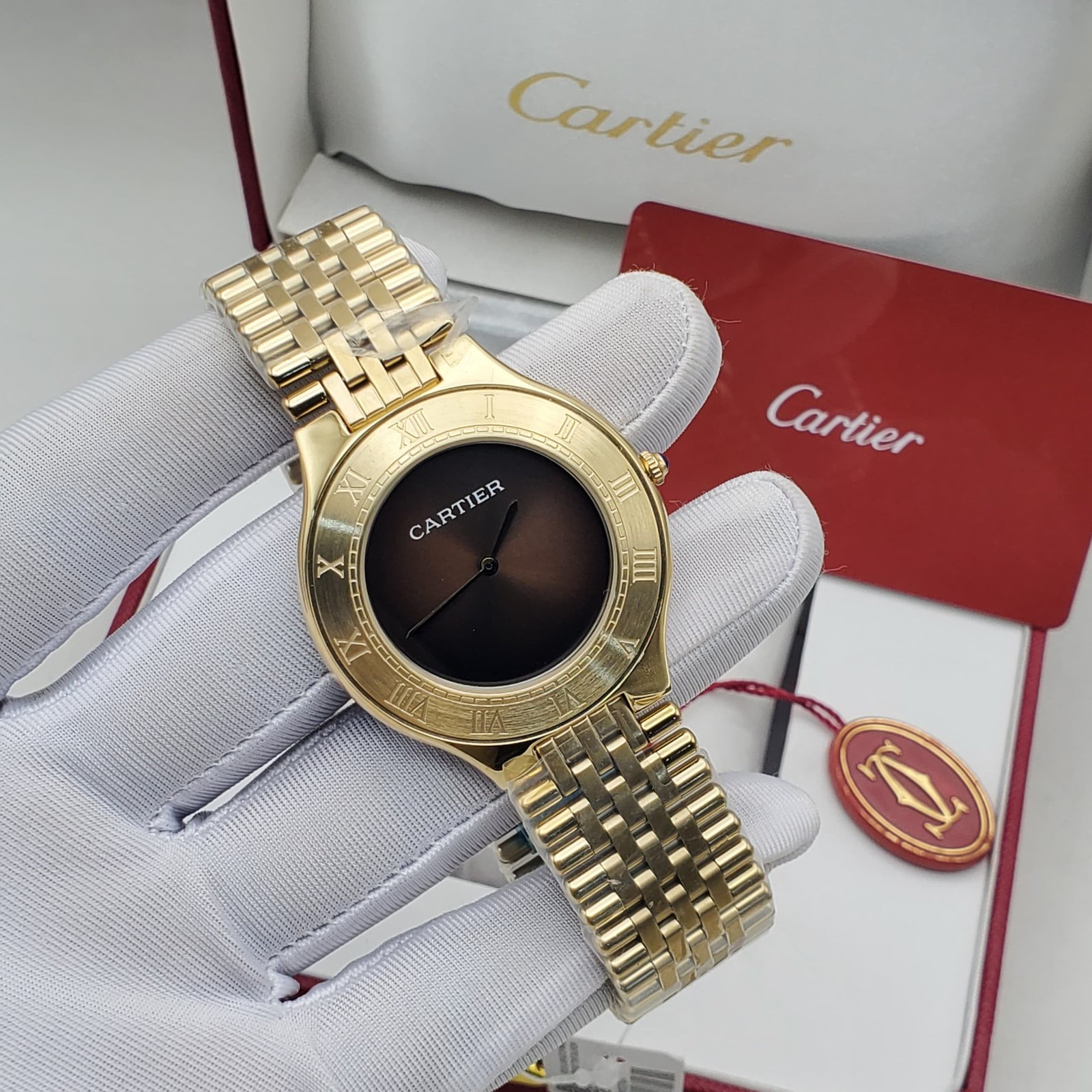 WhatsApp Image 2026-09-06ddd at 8.36.54 PM A gold Cartier watch with a black dial and Roman numerals, displayed on a gloved hand with the brand's packaging in the background.