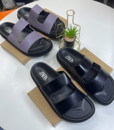 A pair of black and a pair of purple ZAPA slides on brown shoe boxes with a small potted plant in the background.