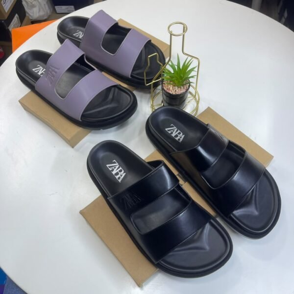 A pair of black and a pair of purple ZAPA slides on brown shoe boxes with a small potted plant in the background.
